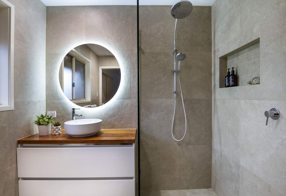 Bathroom renovation completed by Turul Building Services - A modern bathroom with large dark tiles, round backlit mirror, a half-open shower cabin with dual shower head in two sizes